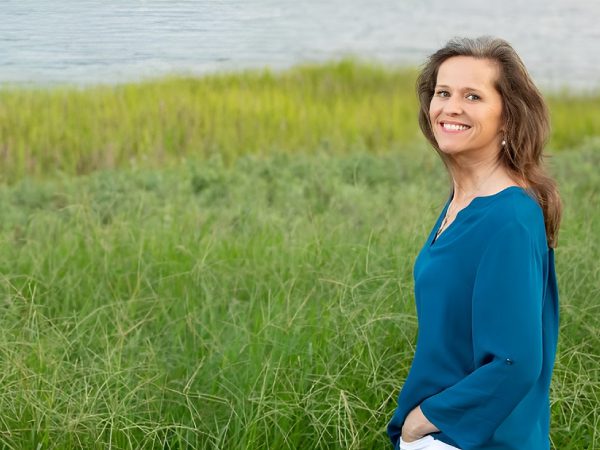 Smiling woman in blue top standing in grassy field near water.
