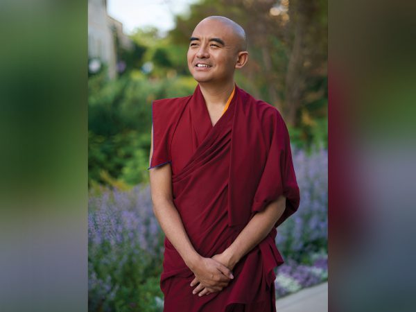 A smiling monk in a red robe standing outdoors surrounded by greenery.