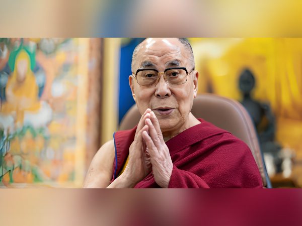 A man in maroon robes with hands clasped, smiling gently.