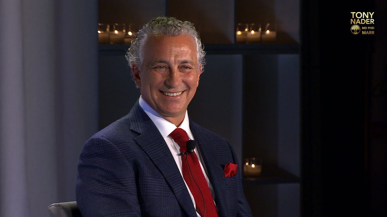A smiling man in a suit with a red tie sitting indoors.