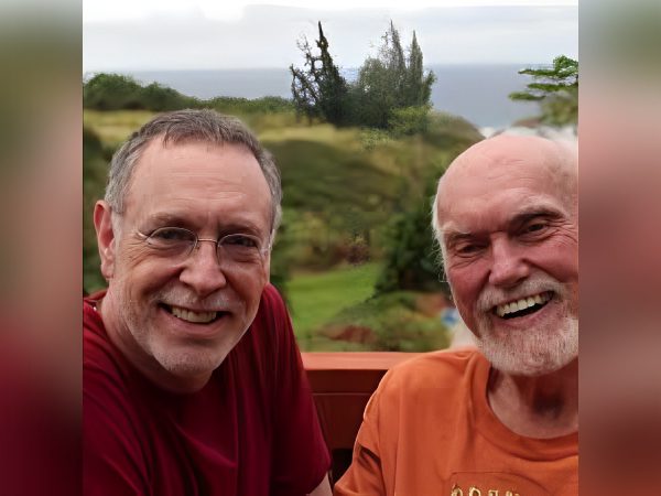 Two older men smiling outdoors with greenery and hills behind them.