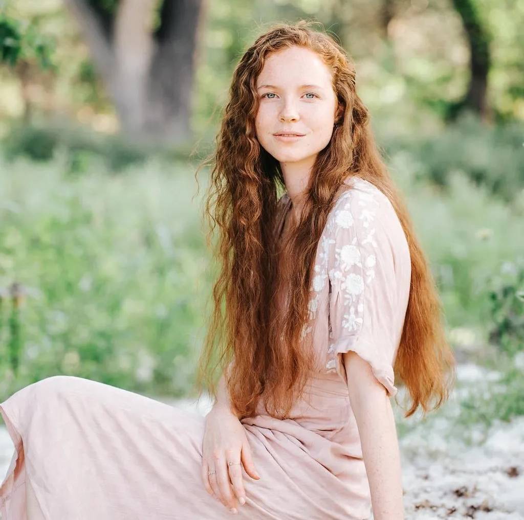 Young woman with long red hair sitting outdoors in a soft pink dress.