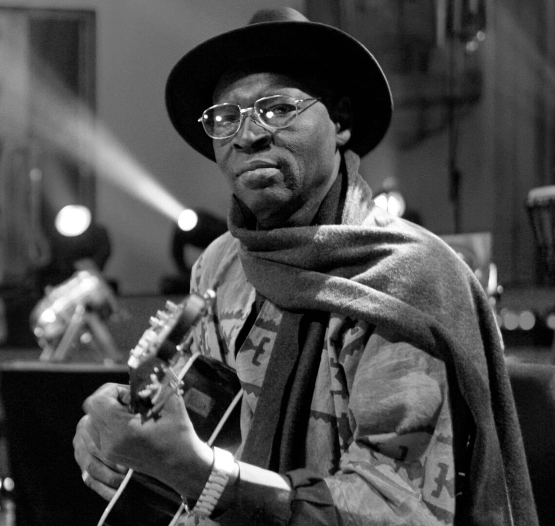 Black and white portrait of a musician playing guitar with a hat and scarf.