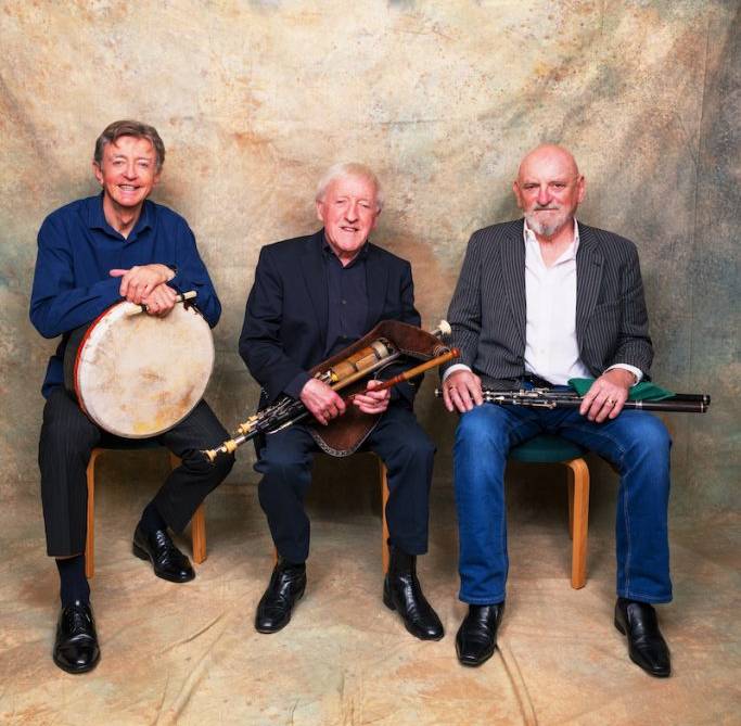 Three elderly men posing with musical instruments against a neutral backdrop.