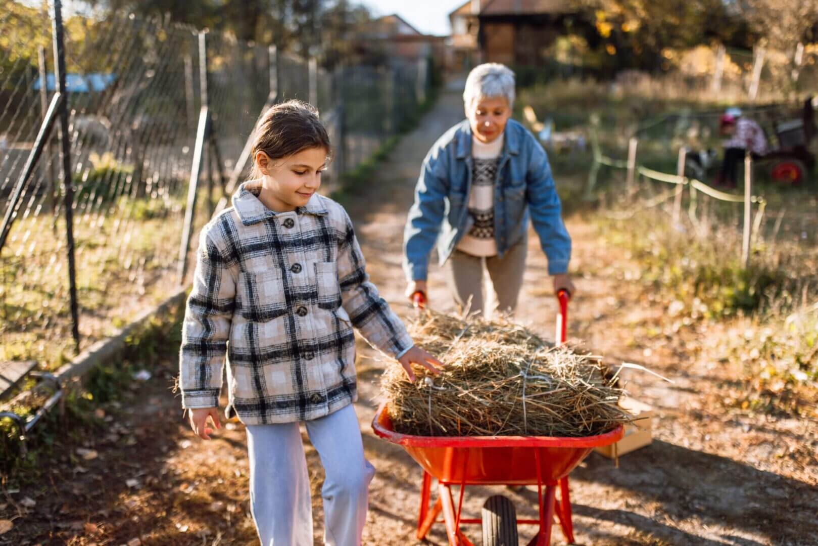 Girl pushing wheelbarrow with elder woman assisting.